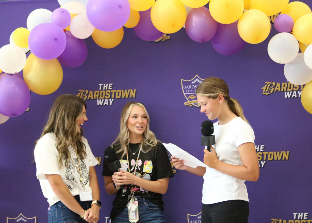 teachers in front of purple backdrop with purple and gold balloons being interviewed by student in white tshirt