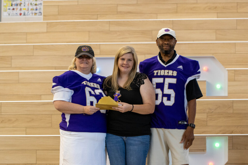 female and male teachers dressed in purple and white athletic attire give award to woman in black shirt and bluejeans