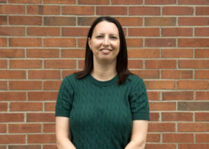 woman with brown hair in a green sweater stands in front of a brick wall
