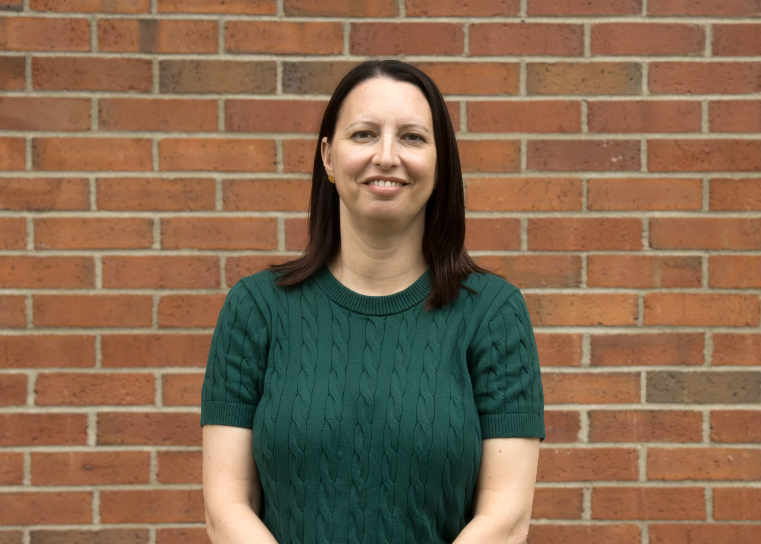 woman with brown hair in a green sweater stands in front of a brick wall